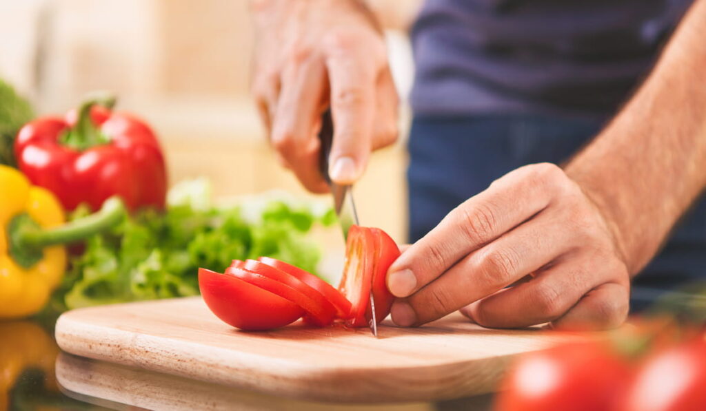 close up of male hand cutting tomato on cutting board at home