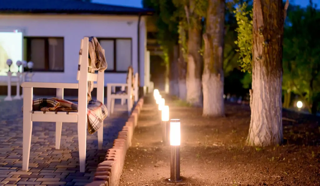 Row of Illuminated Outdoor Lights in Ground Alongside Stone Patio Furnished with Wooden Benches in the yard