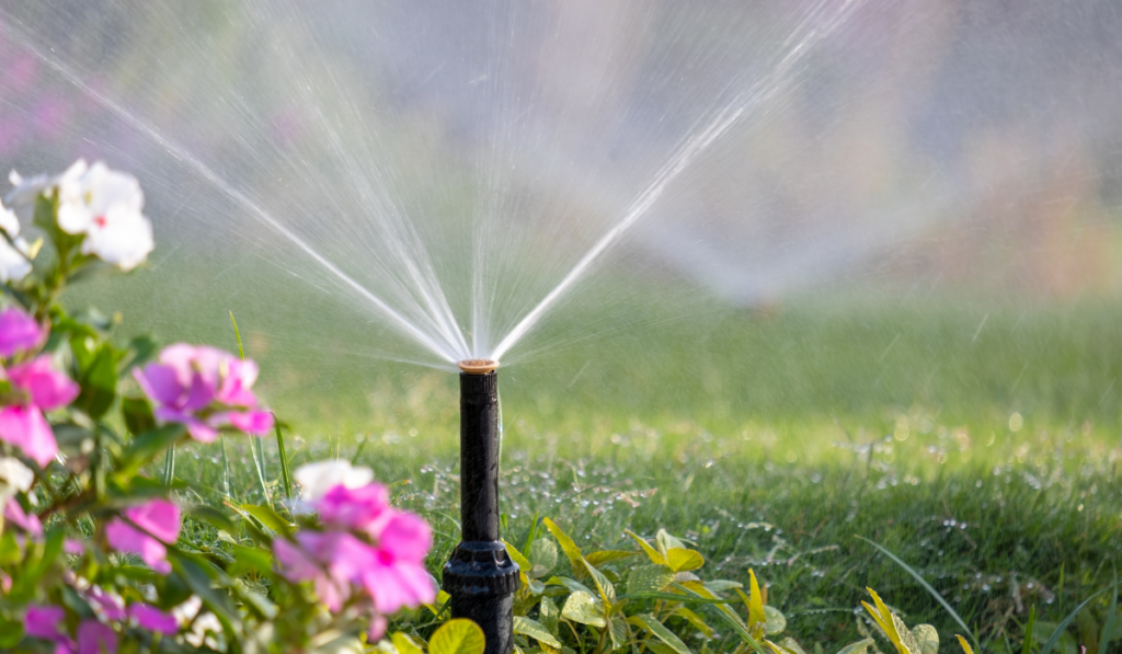 Plastic sprinkler irrigating flower bed on grass lawn with water