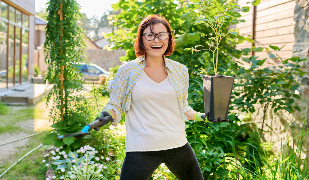 Smiling woman with flowerpot with peony plant and garden shovel
